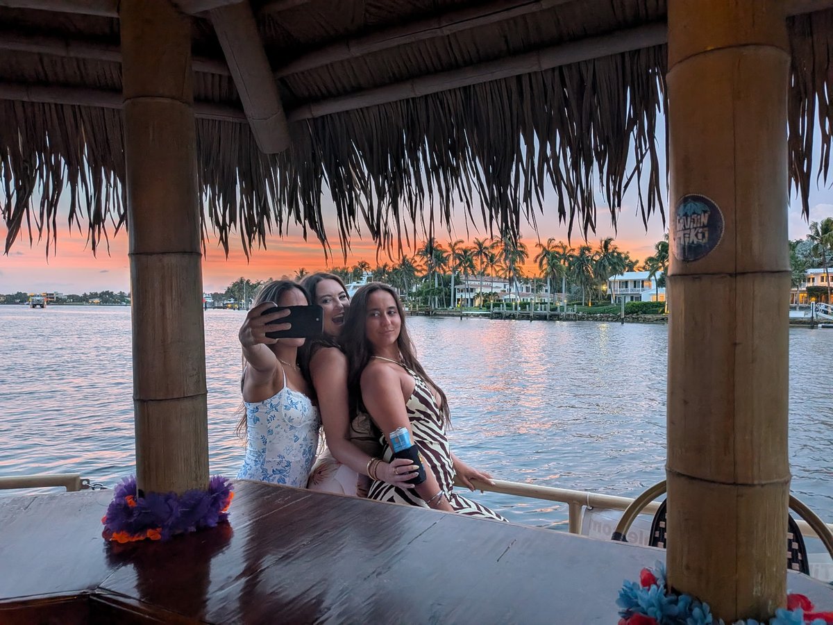 Two women sitting on a wooden pier with thatched roof, posing for a selfie with sunset and palm trees reflected in the water