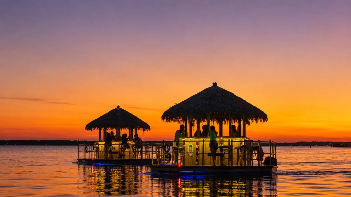 Two illuminated tiki huts on a floating platform in calm water at sunset with orange and purple sky
